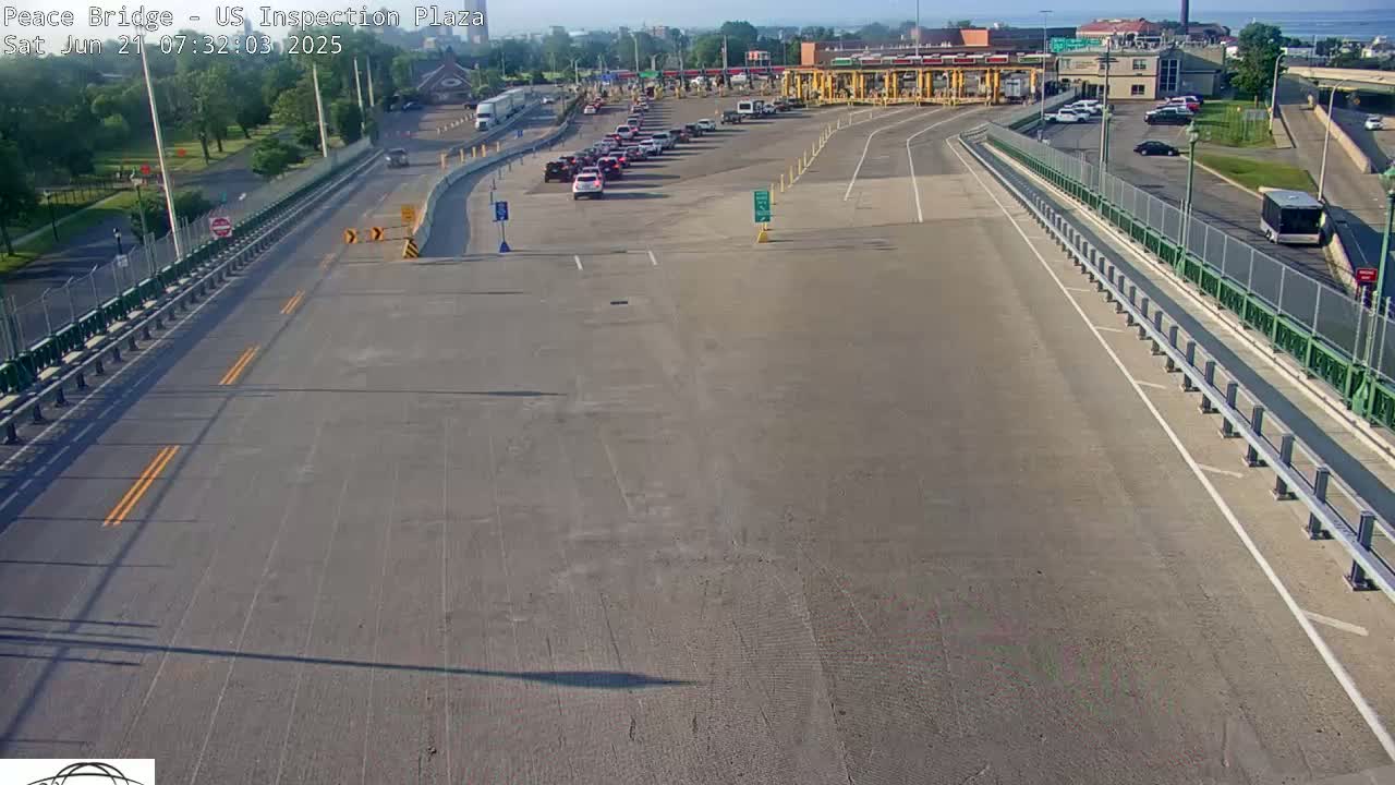 A mostly empty highway approaches a border inspection plaza with a line of cars waiting under a sunny sky.