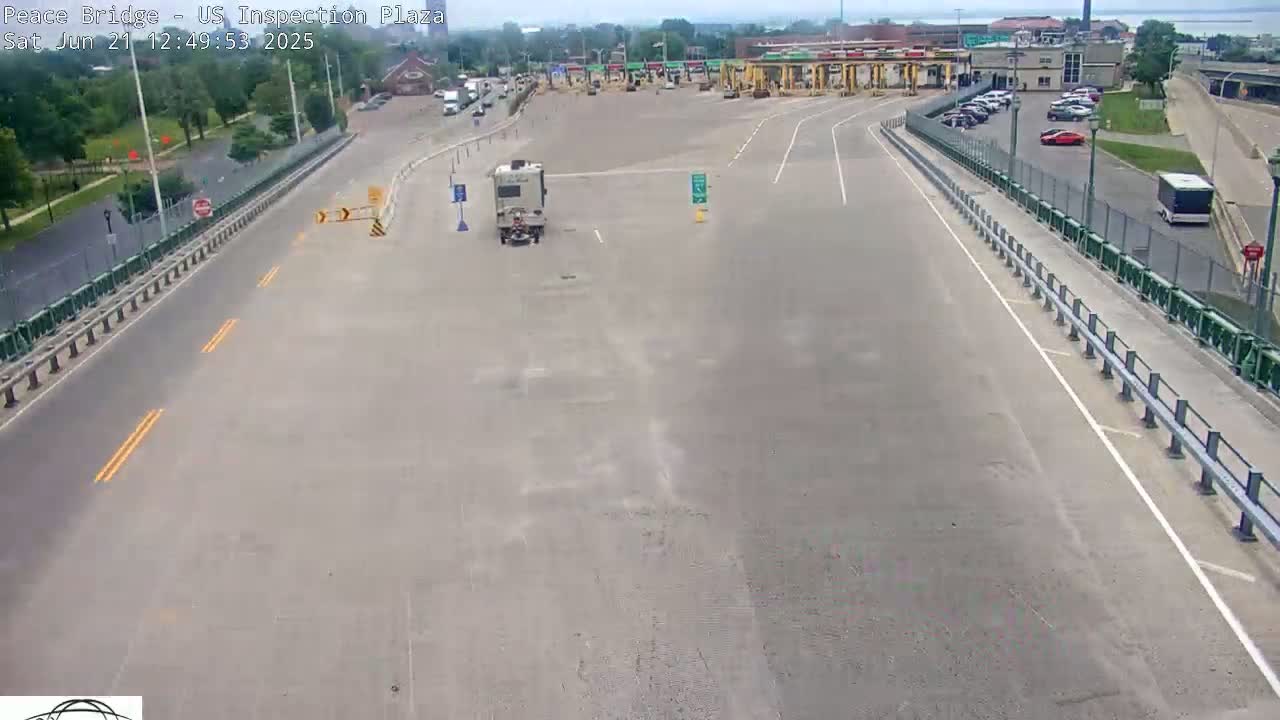 A mostly empty highway approaches a border inspection plaza on a sunny day, with a few vehicles visible in the parking lot and a camper van on the road.