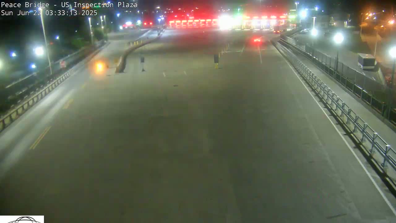 A nighttime, high-angle view shows an empty roadway leading to a border inspection plaza with a few vehicles visible in the distance.