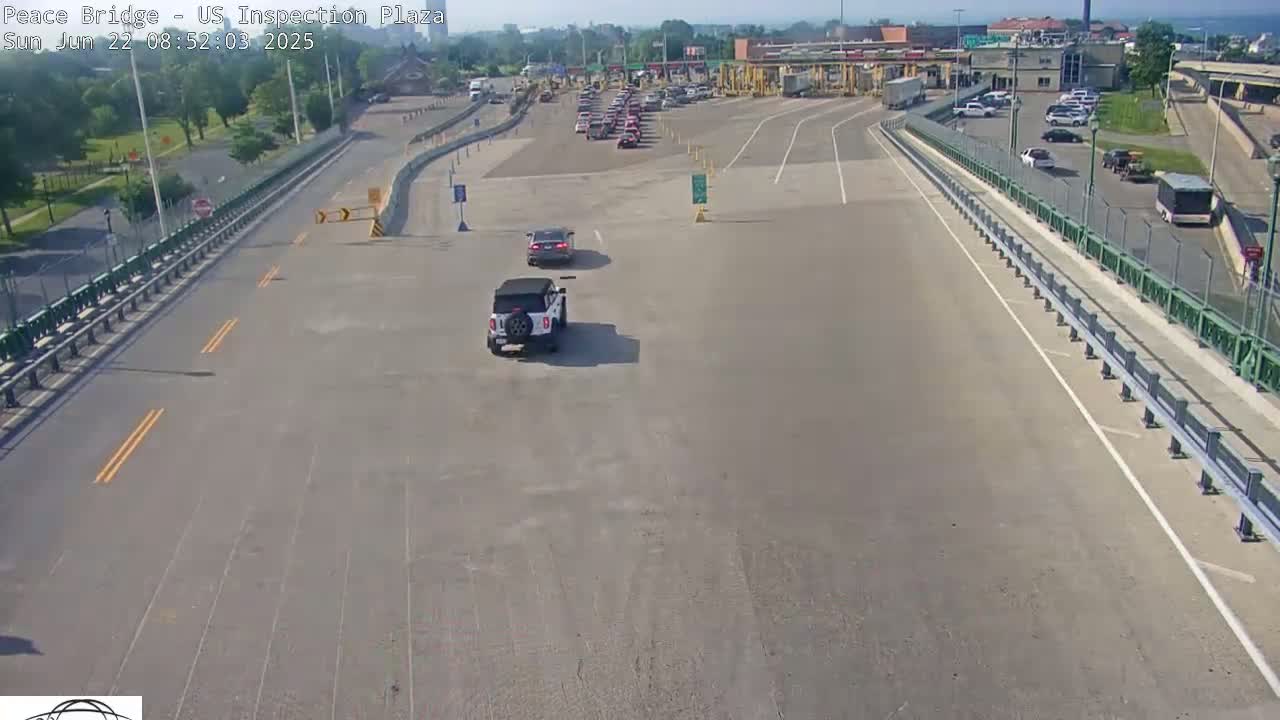 A mostly empty roadway approaches a border crossing inspection plaza on a sunny day, with several vehicles visible in the parking lot and on the road.