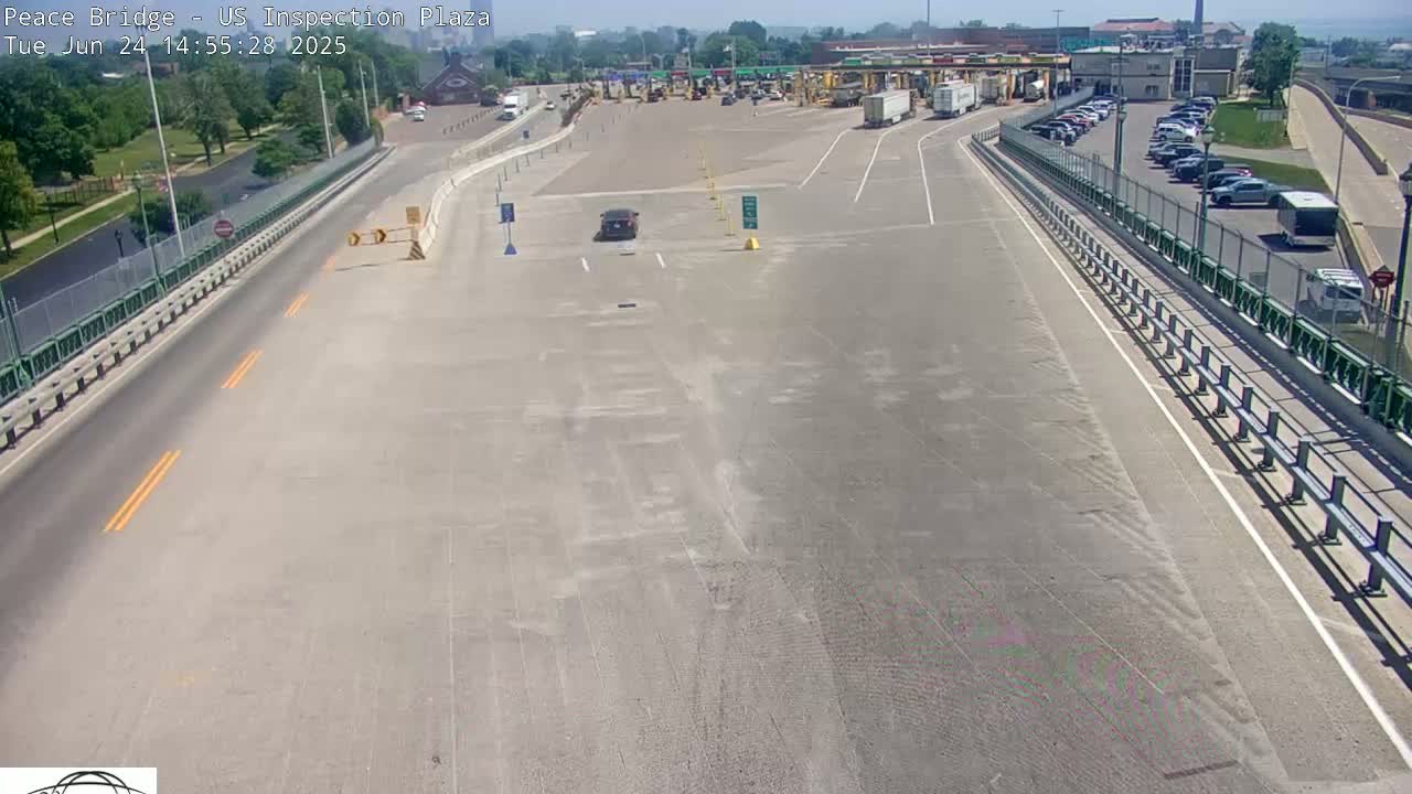 A mostly empty highway approaches a border inspection plaza on a sunny day, with a few cars and trucks visible in the parking lot and a single vehicle on the roadway.