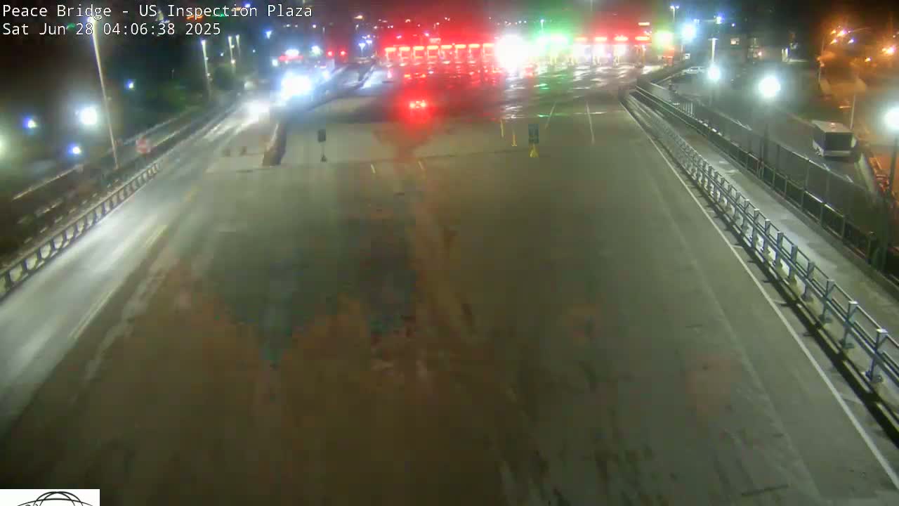 A nighttime, wet road leads to a border crossing inspection plaza with a few vehicles present.