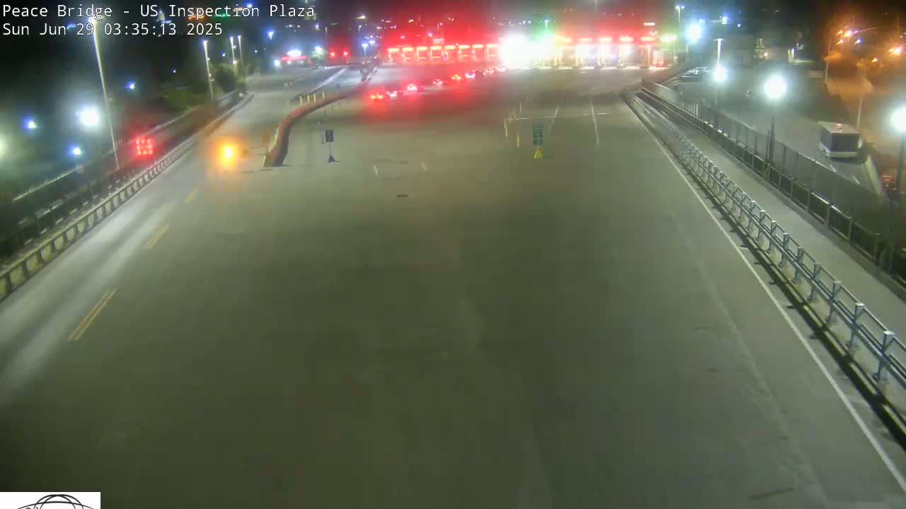 A nighttime overhead view shows a mostly empty roadway leading to a border crossing inspection plaza with a few vehicles present.