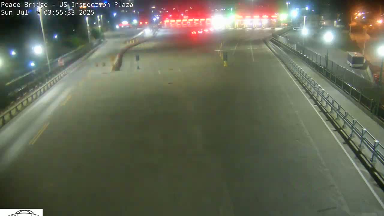 A mostly empty roadway at night leads to a border crossing inspection plaza with a few vehicles present.