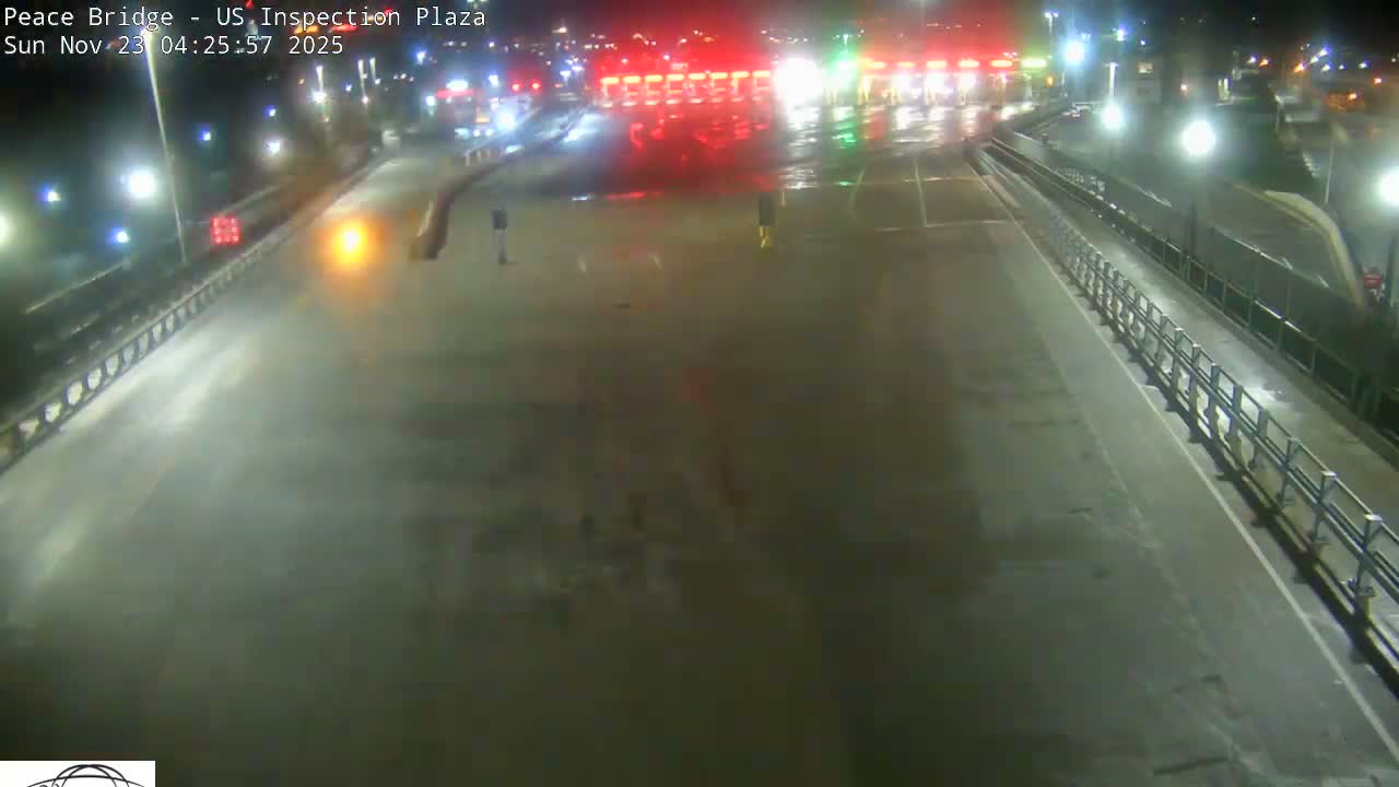 The image shows a wet highway or bridge at night, illuminated by numerous streetlights and bright red and green lights from a structure in the distance, with a few figures visible on the pavement.