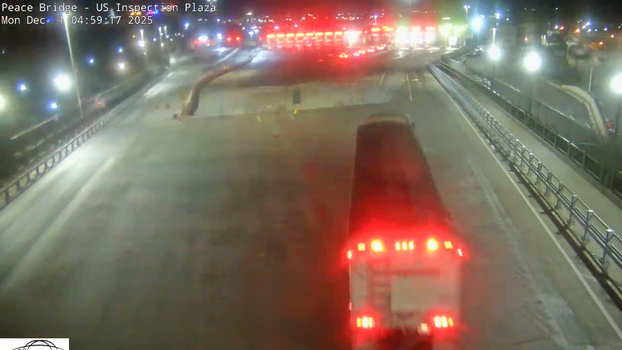 A large truck with bright red taillights drives on a multi-lane bridge or highway under clear night conditions, approaching an illuminated inspection plaza with many red lights visible in the distance.