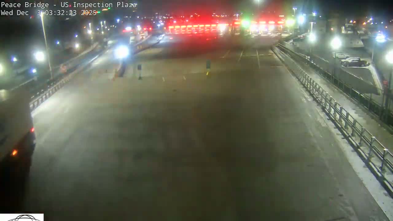 A wet, multi-lane road at night leads to an illuminated inspection plaza with red and green lights, reflecting in the damp pavement.