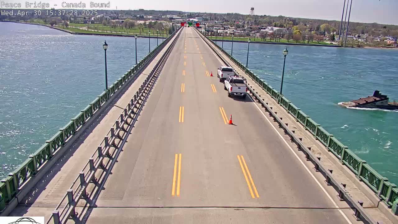 A bridge over a choppy body of water on a sunny day has two white vehicles and traffic cones on its roadway.