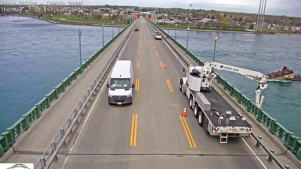 A bridge over blue water shows several vehicles on the road, including a white van and a flatbed truck with a crane, under a mostly sunny sky.