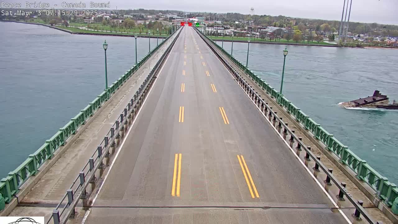 A mostly empty, wet roadway spans a bridge over water, with a few vehicles visible in the distance under an overcast sky.