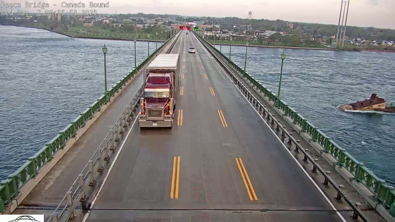 A long bridge over water shows a semi-truck and several cars driving on a wet road, with a partially submerged vessel visible in the water.