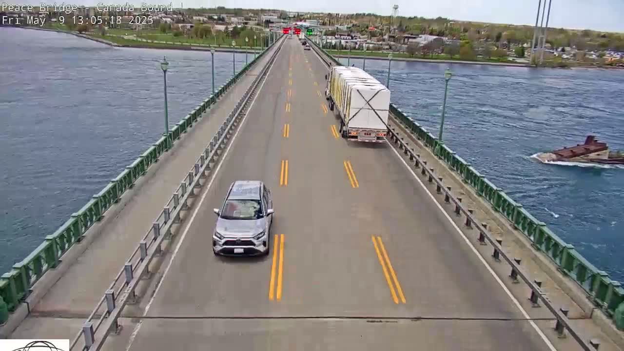 A silver SUV and a large white-walled semi-truck are driving on a bridge over a body of water on a partly sunny day.
