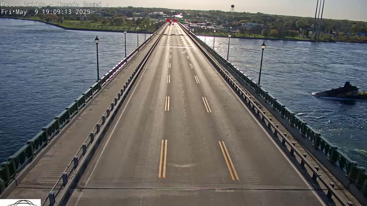 A long, mostly empty bridge road stretches across a body of water under a sunny sky, with a single vessel visible in the water.