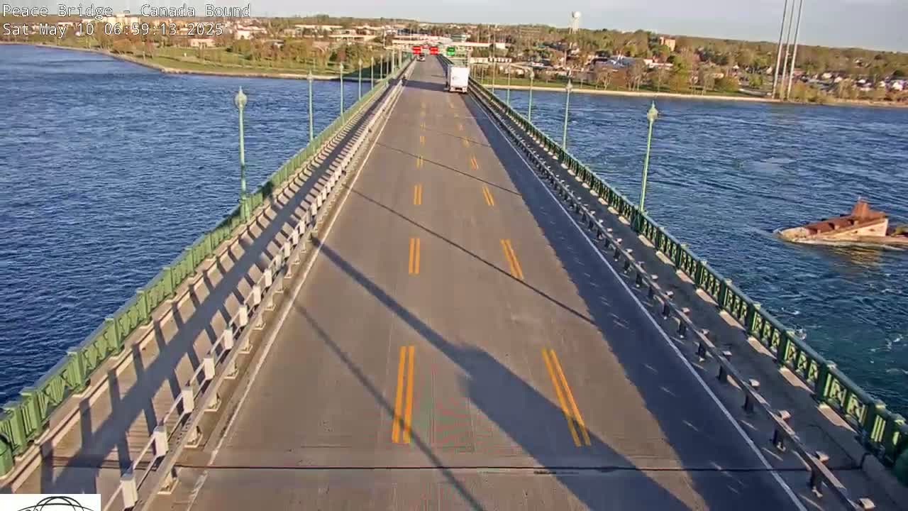 A bridge road with a few vehicles is seen, with calm blue water on either side under a mostly sunny sky.