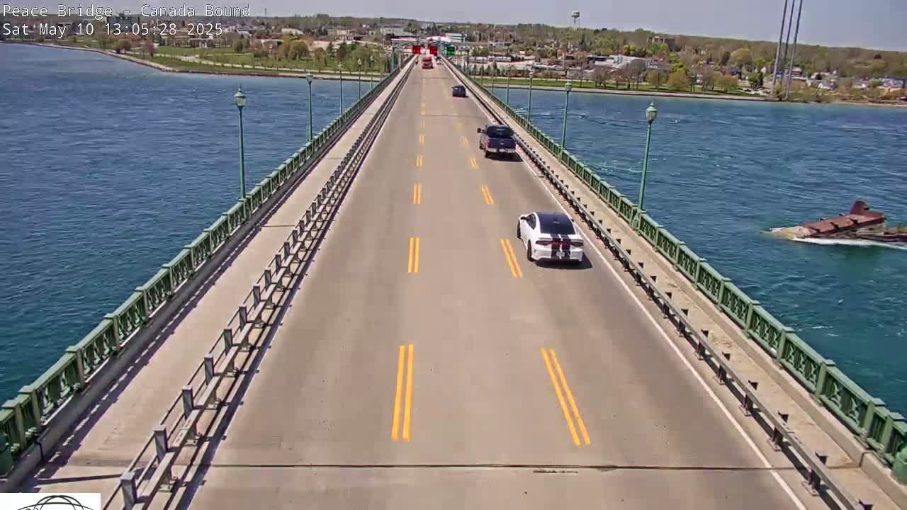 A two-lane bridge over blue water on a sunny day has several cars driving on it, with a barge visible in the water below.