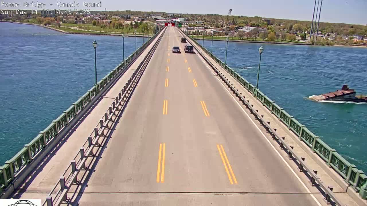 A two-lane bridge spans a body of water on a sunny day, with a few cars driving across and a sunken vessel visible in the water to the right.