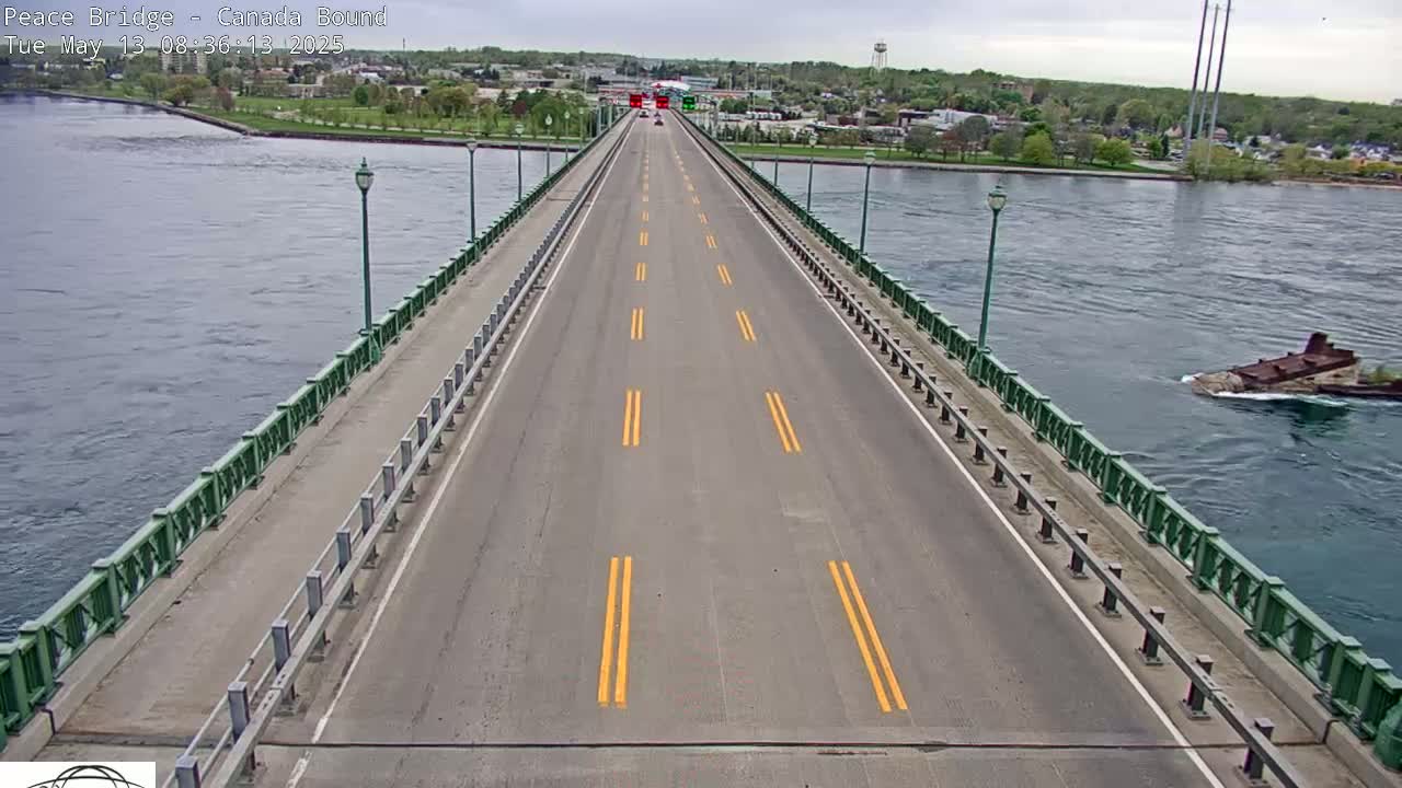 A long bridge roadway with a few vehicles stretches across a body of water under an overcast sky, with a partially submerged vessel visible in the water to the right.