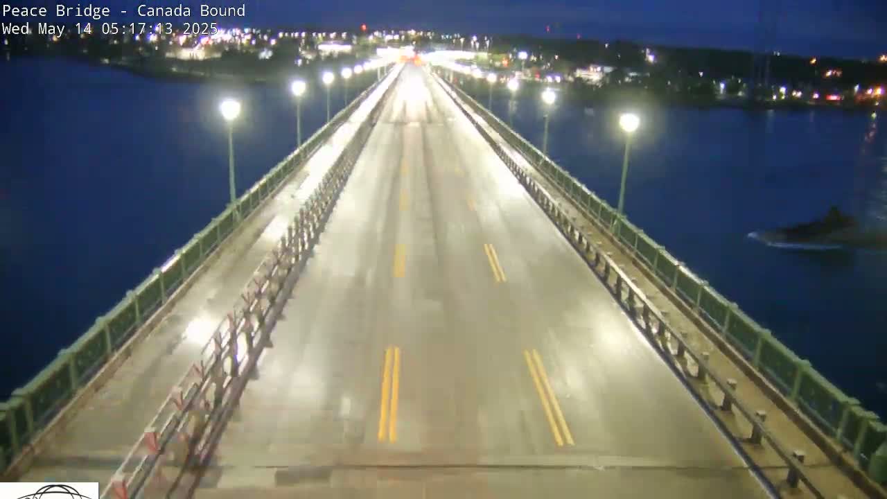 A wet, two-lane bridge is lit at night by lamps on either side, with a few vehicles visible in the distance and dark water below.