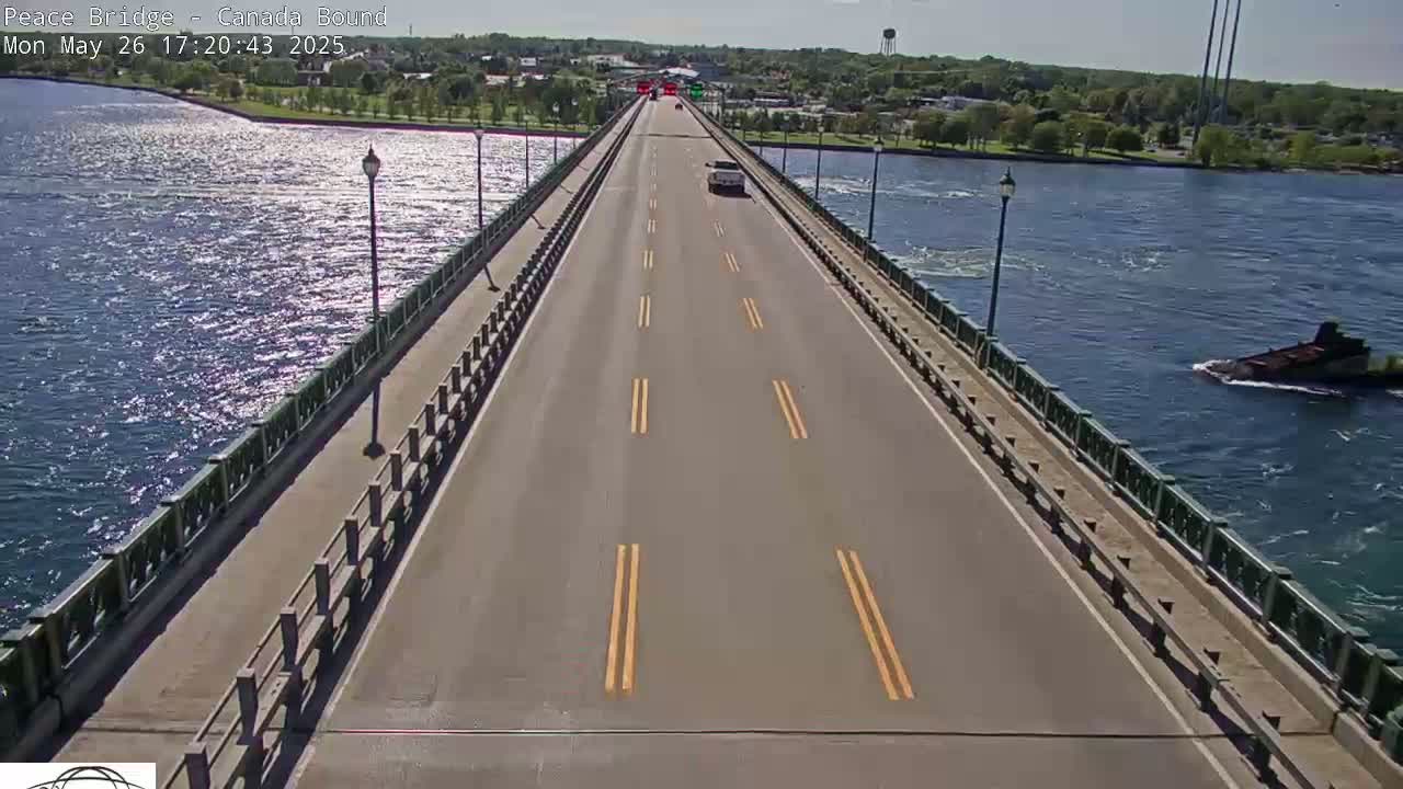 A bridge with a few cars driving on it stretches over a body of water on a sunny day; a barge is visible in the water to the right.