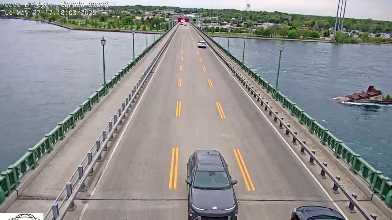 A bridge roadway with several cars, bordered by green railings, spans a body of water under a partly cloudy sky.