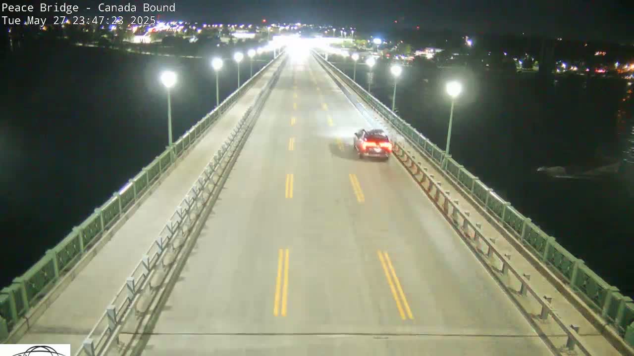 A single car drives at night across a bridge over dark water, with lights illuminating the roadway.