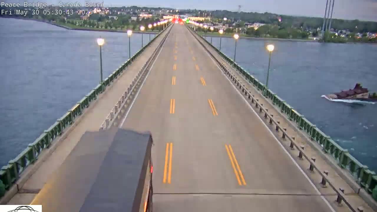 A long bridge stretches over calm water under an overcast sky, with a vehicle driving on it and a barge in the water below.
