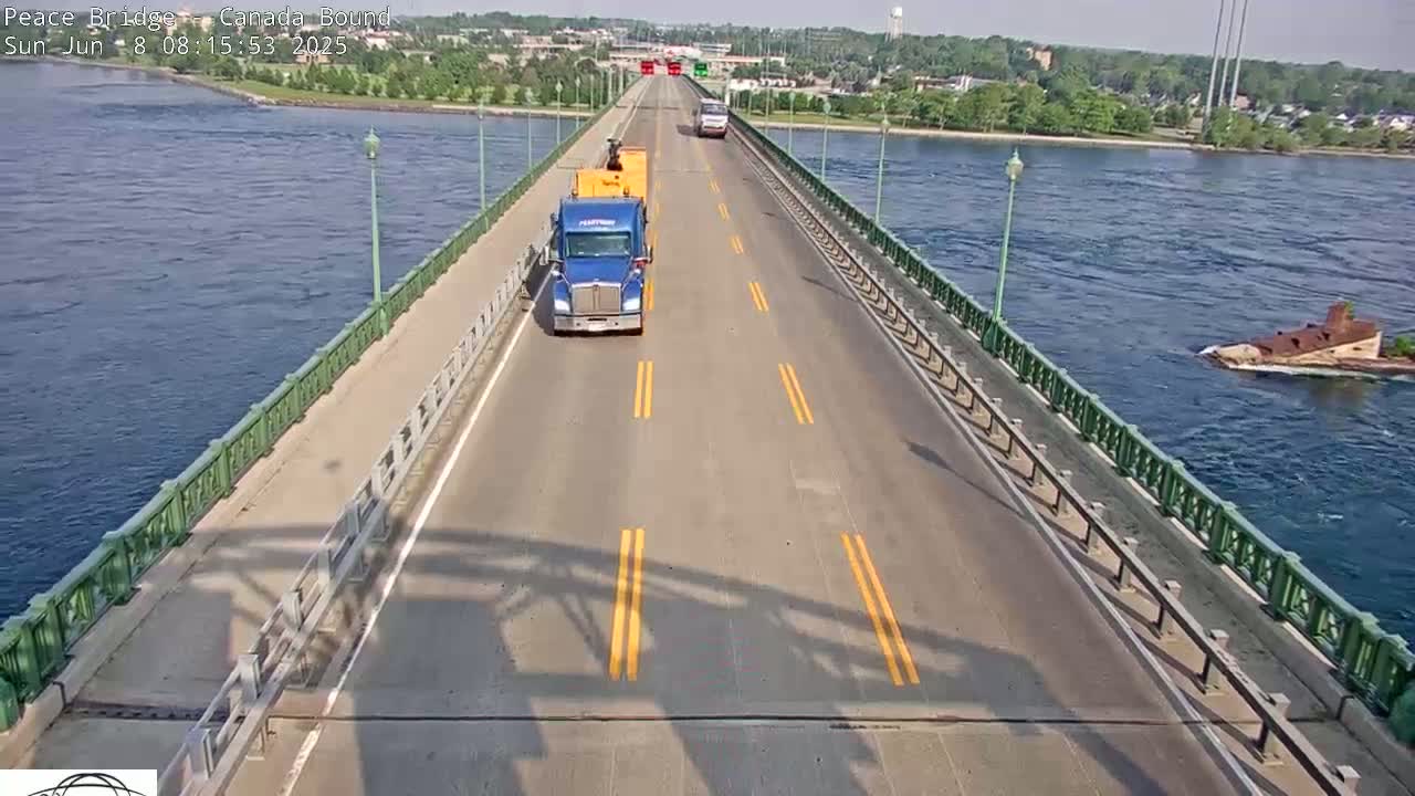 A sunny day shows several vehicles driving on a bridge over water, with a barge visible in the water.