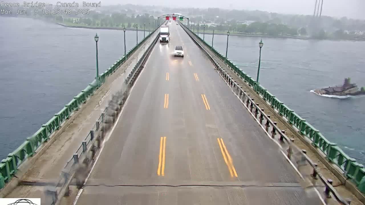 A wet, two-lane bridge over water carries a few vehicles in rainy weather; a barge is visible in the water below.