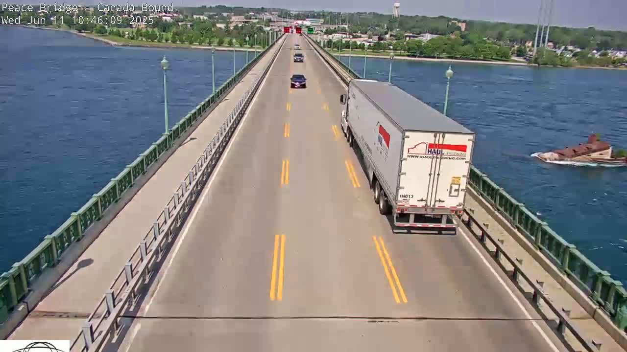 A long bridge over water on a sunny day with several vehicles traveling on it, including a large semi-truck, and a small vessel in the water beside the bridge.
