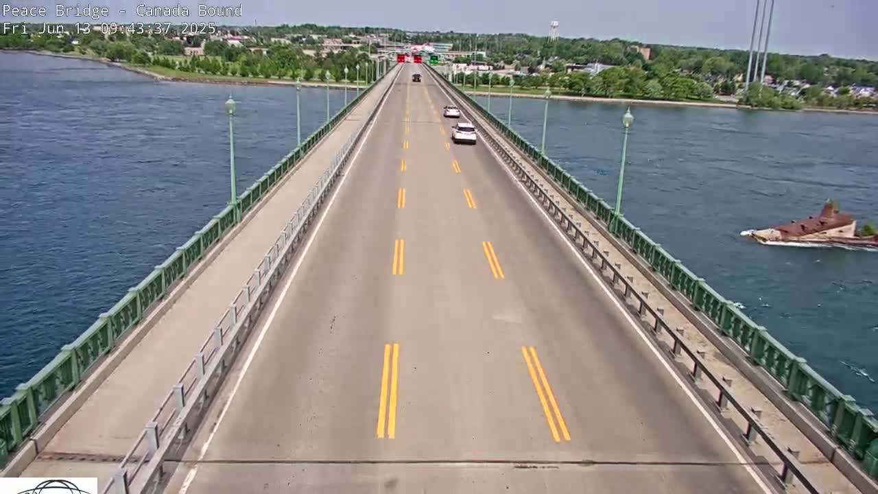 A bridge roadway with a few cars on it stretches across a body of water on a sunny day, with a partially submerged structure visible in the water to the right.