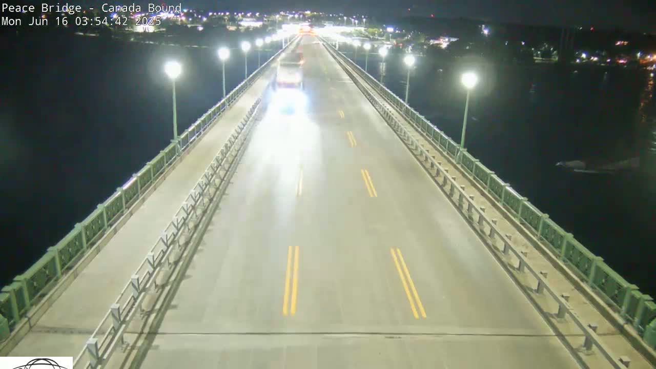 A nighttime, overhead view shows a bridge road with a few cars driving on it, bordered by railings over dark water.