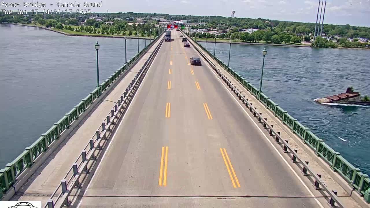 A bridge with several cars driving on it stretches across a body of water on a sunny day, with green railings and land visible on either side.