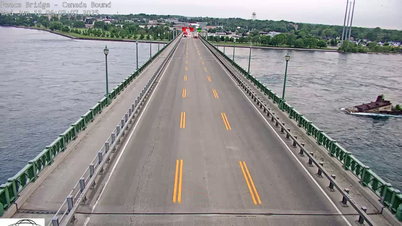 A long bridge road with yellow lines stretches across a body of water on an overcast day, with a small vessel visible in the water to the right.