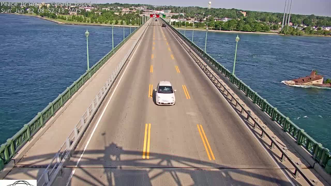 A white car drives on a bridge over blue water on a sunny day, with another vehicle visible in the distance.