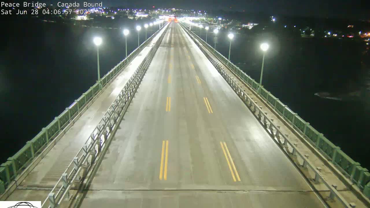A wet, nighttime view of a bridge road with few cars and bright lights on either side.