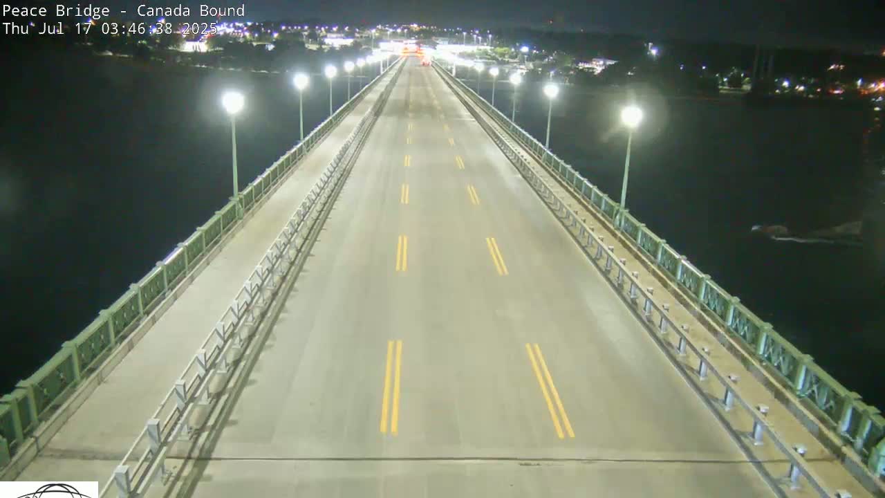 A mostly empty roadway on a bridge at night, illuminated by streetlights, overlooks dark water.