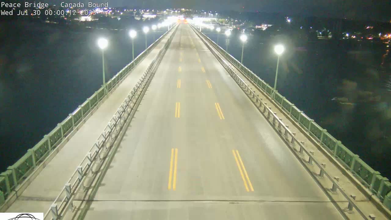 A mostly empty roadway on a bridge at night, illuminated by streetlights, overlooks dark water.