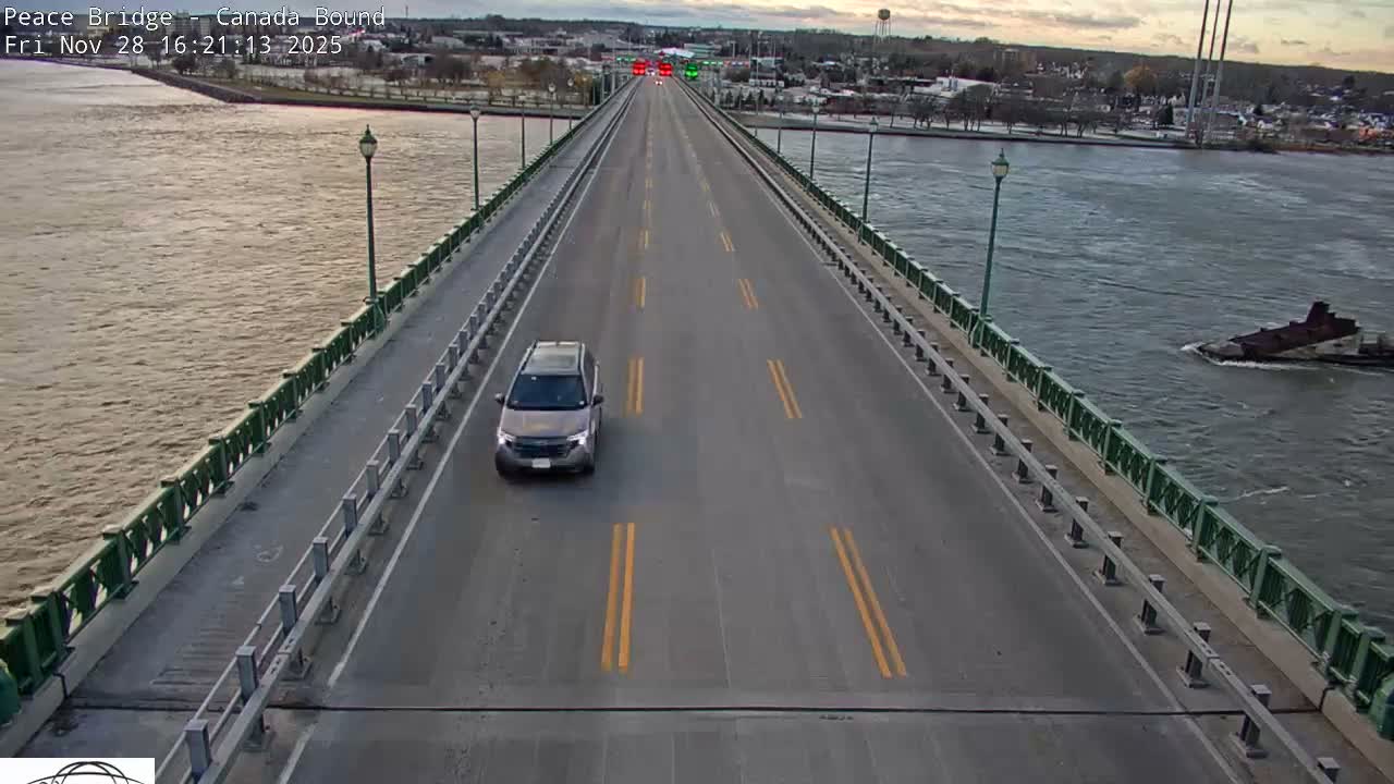 A grey SUV travels over a long, multi-lane bridge spanning a wide river with a barge visible on the right, under an overcast sky with faint sunset hues over the distant city.