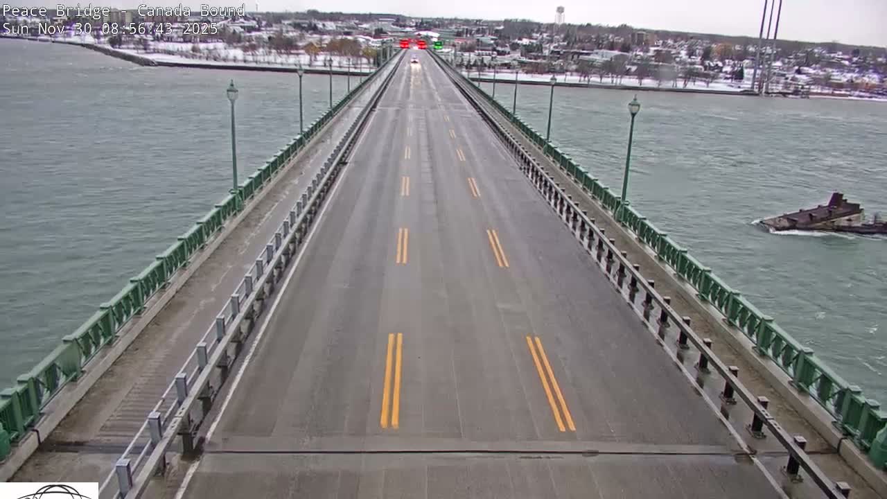 A grey SUV travels over a long, multi-lane bridge spanning a wide river with a barge visible on the right, under an overcast sky with faint sunset hues over the distant city.