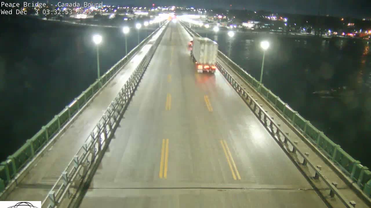 A truck drives over a wet, streetlight-illuminated bridge at night, with distant city lights reflecting on the water below.