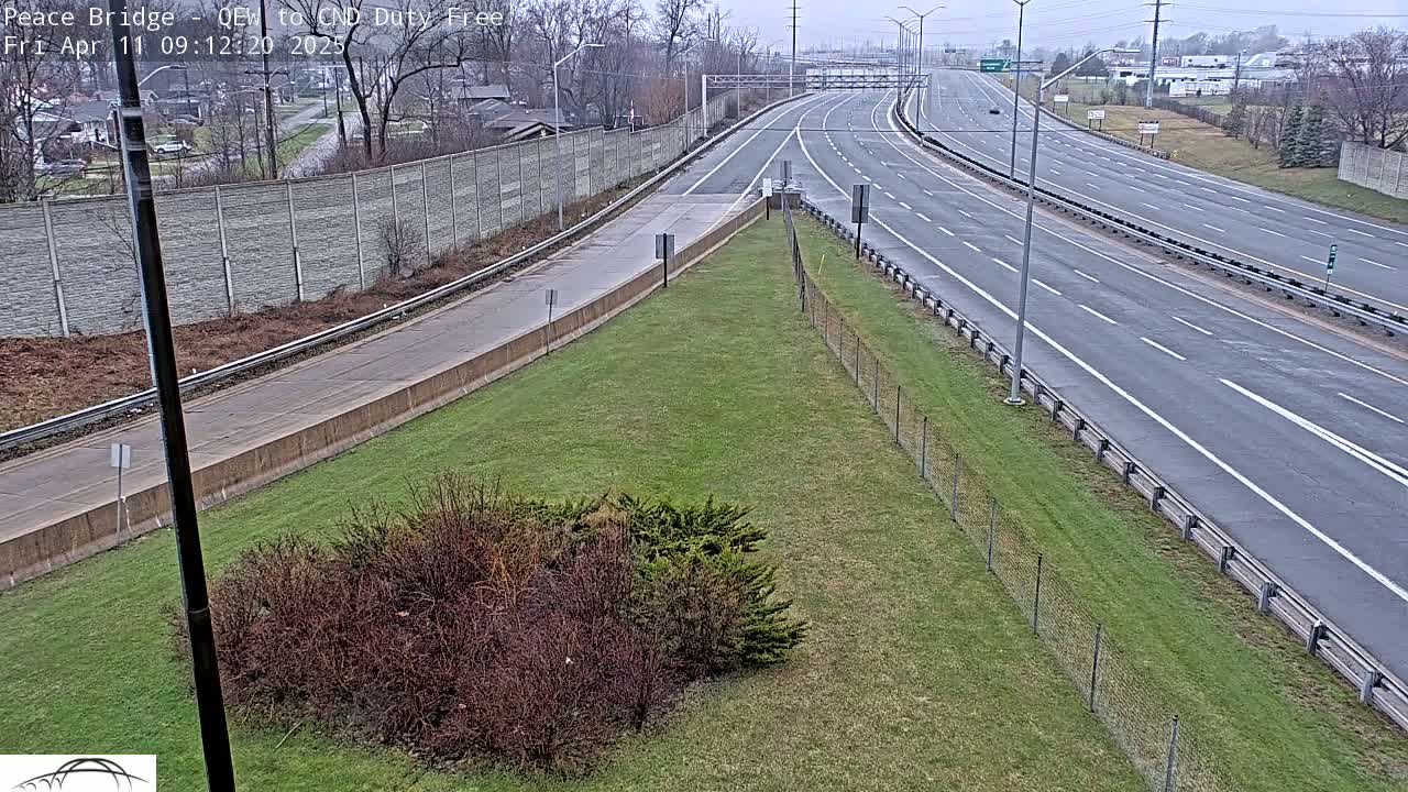 A multi-lane highway stretches into the distance on an overcast day, bordered by a grassy area with sparse shrubs and a chain-link fence.