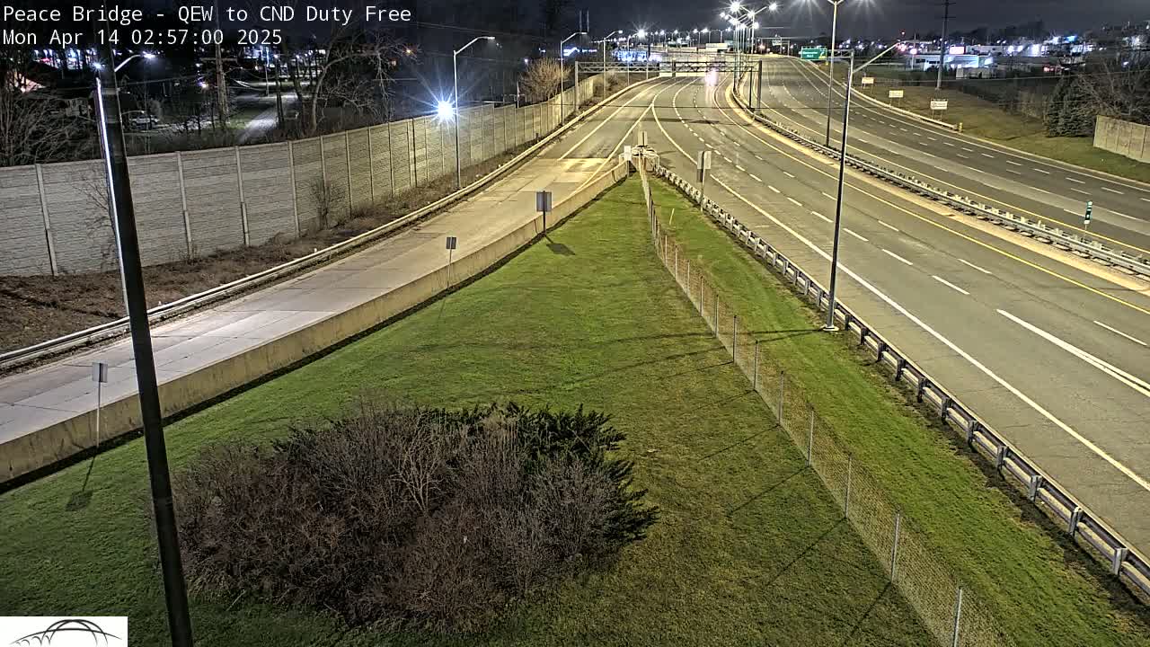 A nighttime view of a highway interchange with sparse traffic, bordered by a grassy area and a chain-link fence.