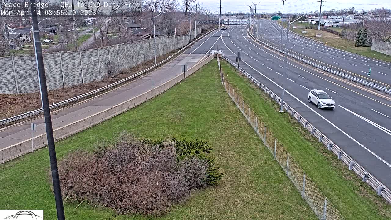 A mostly clear day reveals a multi-lane highway with sparse traffic, alongside a grassy area bordered by a concrete wall and chain-link fence.