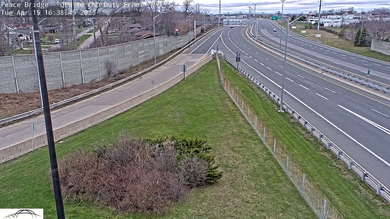 A partly cloudy day reveals a multi-lane highway with sparse traffic, bordered by a grassy area and a concrete wall separating it from residential buildings.