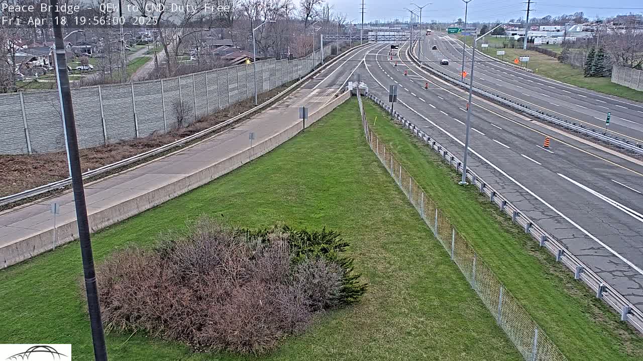 A mostly empty multi-lane highway with sparse traffic on a partly cloudy day, bordered by a chain link fence and grassy areas.