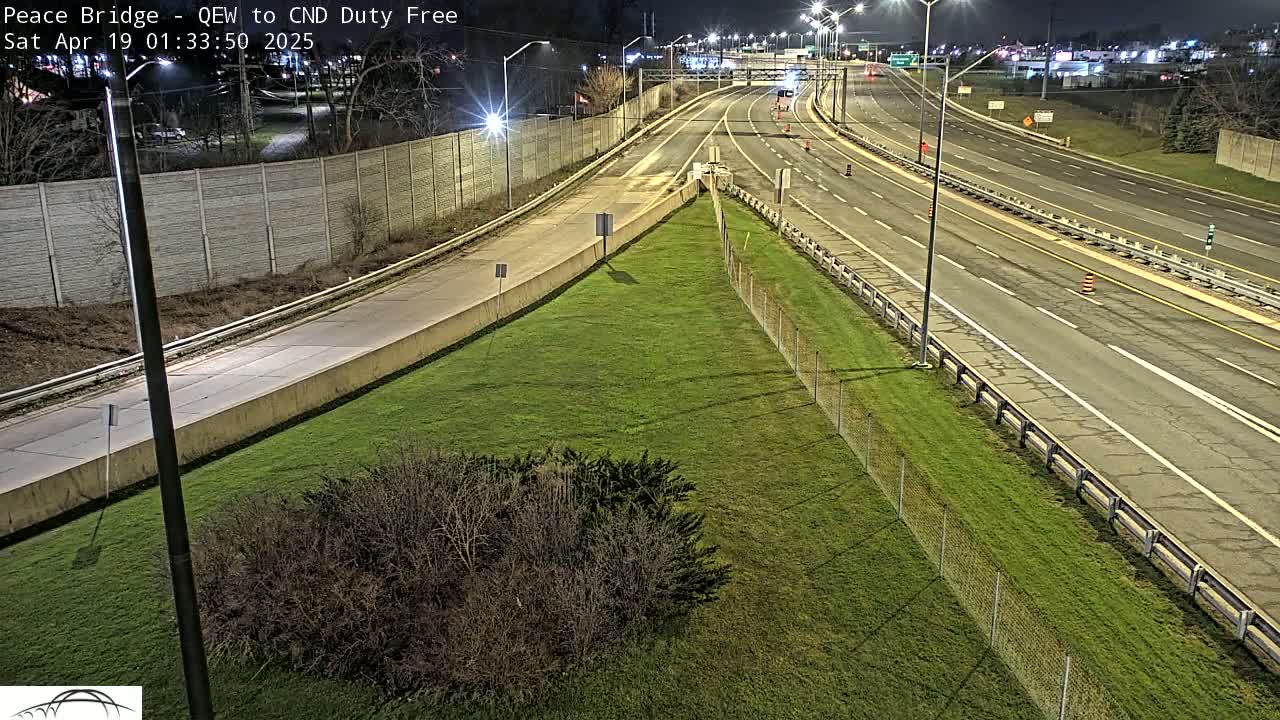 A nighttime, overhead view shows a multi-lane highway with sparse traffic, bordered by a grassy area with a chain-link fence.