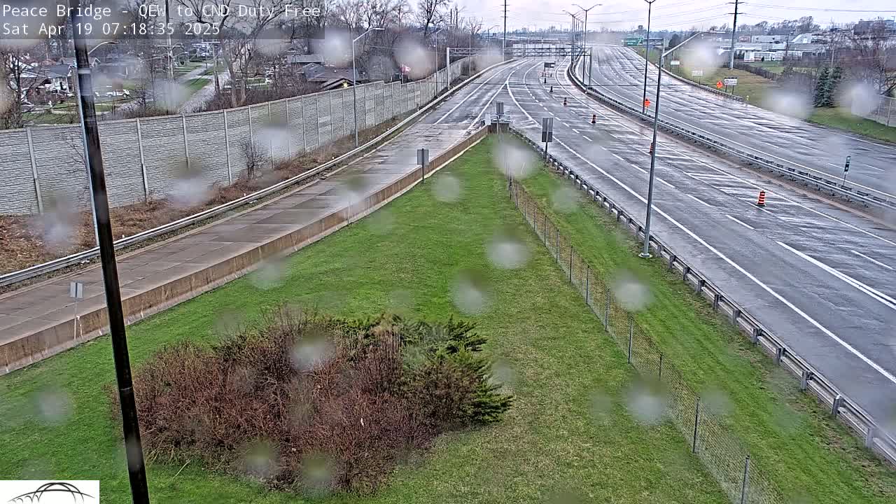 A wet highway merges onto a larger highway, with grassy areas and a fence separating the roads from surrounding shrubbery and a distant wall.
