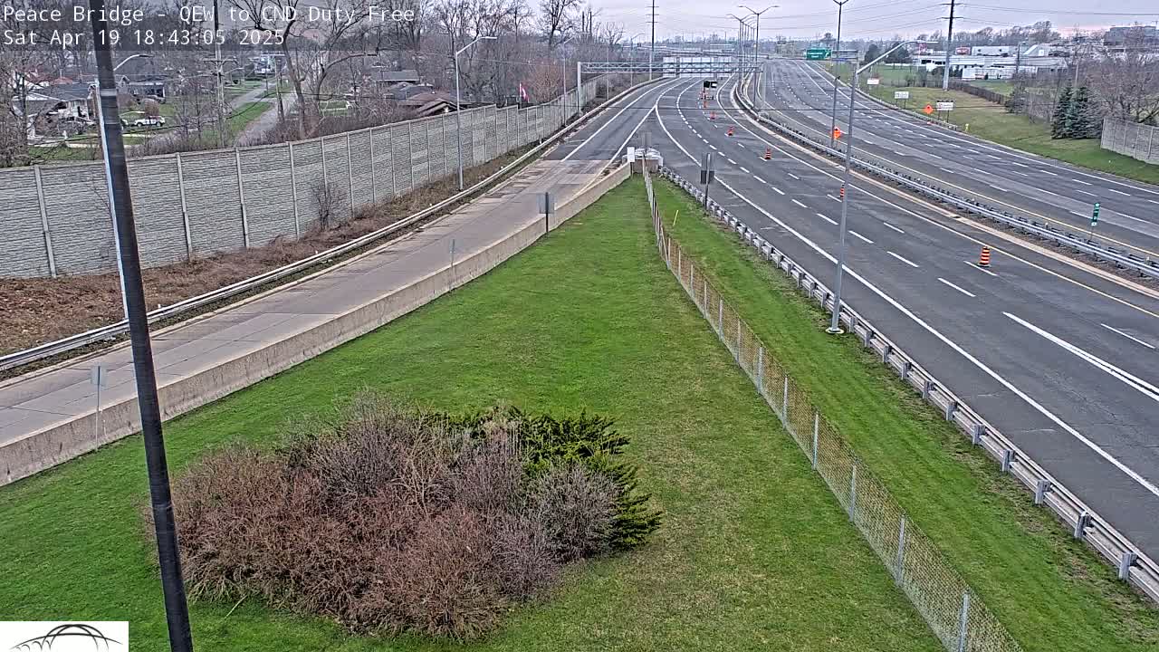 A mostly empty highway stretches into the distance under an overcast sky, bordered by grassy areas and a chain-link fence.