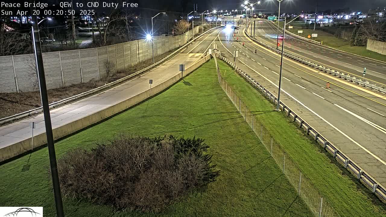 A nighttime view of a highway interchange with sparse traffic, surrounded by grassy areas and a chain-link fence.