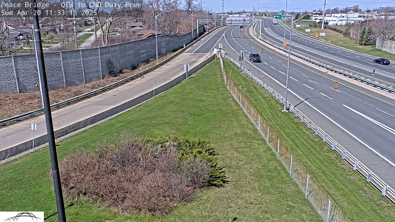 A highway scene on a sunny day shows a few cars traveling on a multi-lane roadway bordered by grassy areas and chain-link fences.